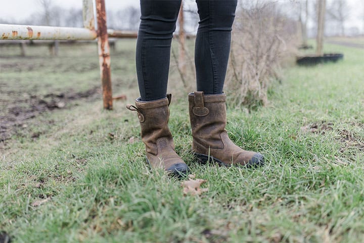 Person standing on green grass wearing brown, muddy boots and black pants; background shows a rusty metal fence and out-of-focus natural scenery.