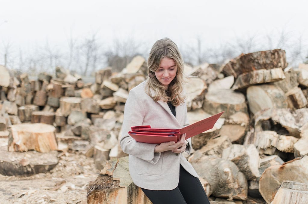 A woman with blonde hair in a light coat sits on a log outdoors, holding and reading from a red folder. Piles of chopped firewood are stacked in the background on a cloudy day.