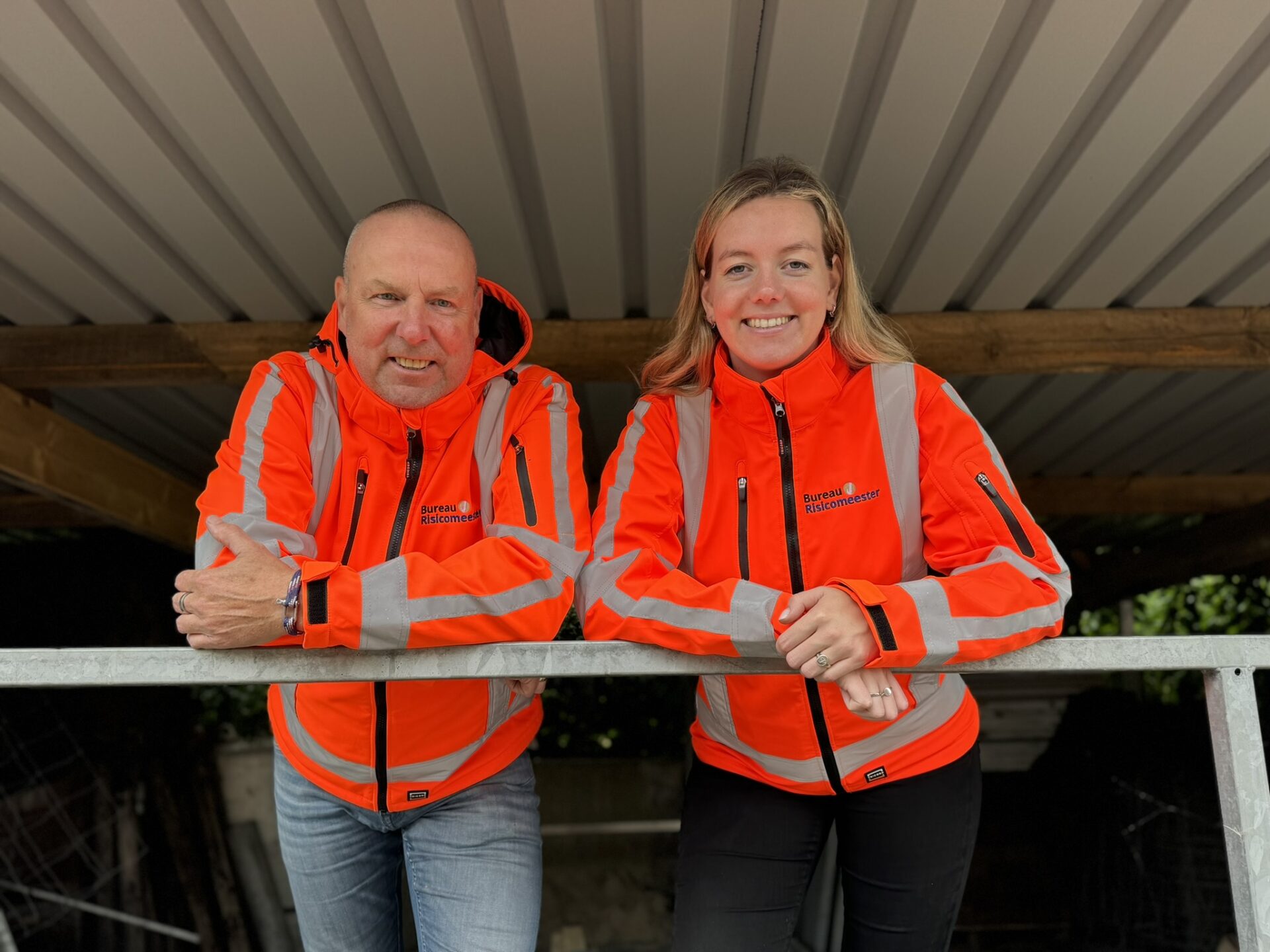 Two people wearing bright orange and gray high-visibility jackets stand side by side, leaning on a metal railing under a corrugated roof, smiling at the camera.