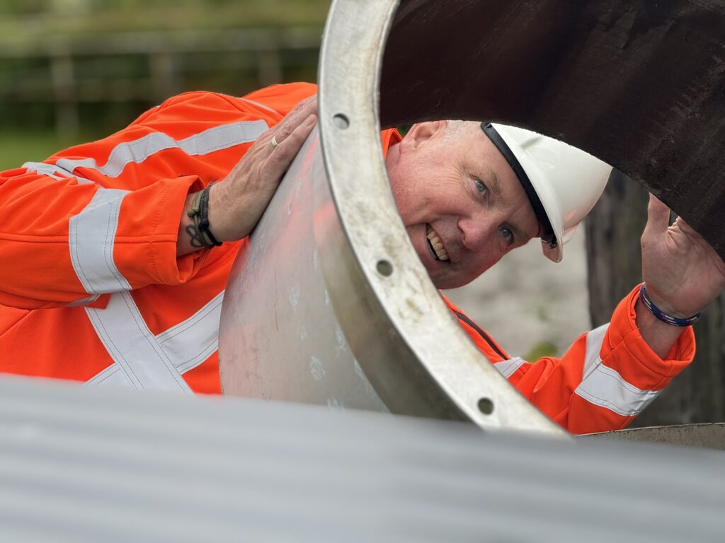 A man in a white hard hat and orange high-visibility jacket smiles while peeking through a large metal pipe outdoors.