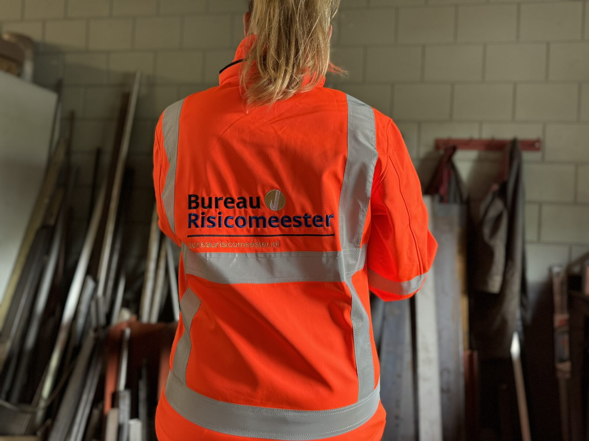 A person wearing a bright orange safety jacket with reflective stripes stands with their back to the camera. The jacket reads Bureau Risicomeester in black letters. The background shows metal sheets and a tiled wall.